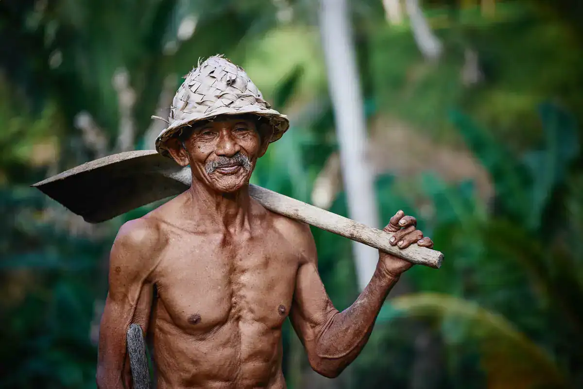 Elderly man with a woven hat carrying a hoe in a lush green environment.
