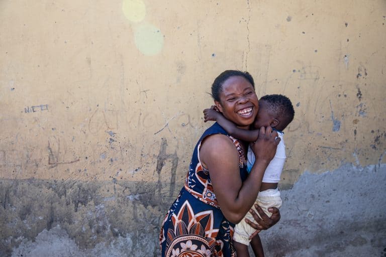 A joyful woman with a big smile, hugging a young child outdoors.