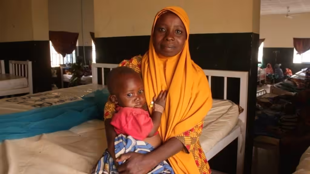 A woman with a headscarf smile to the camera as she holds a young child in a hospital room.