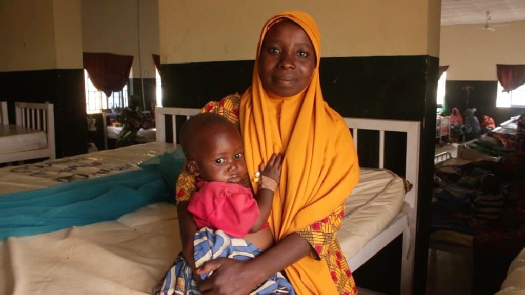 A woman with a headscarf smile to the camera as she holds a young child in a hospital room.