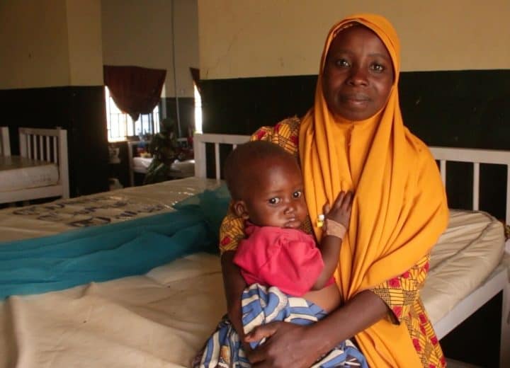 A woman with a headscarf smile to the camera as she holds a young child in a hospital room.