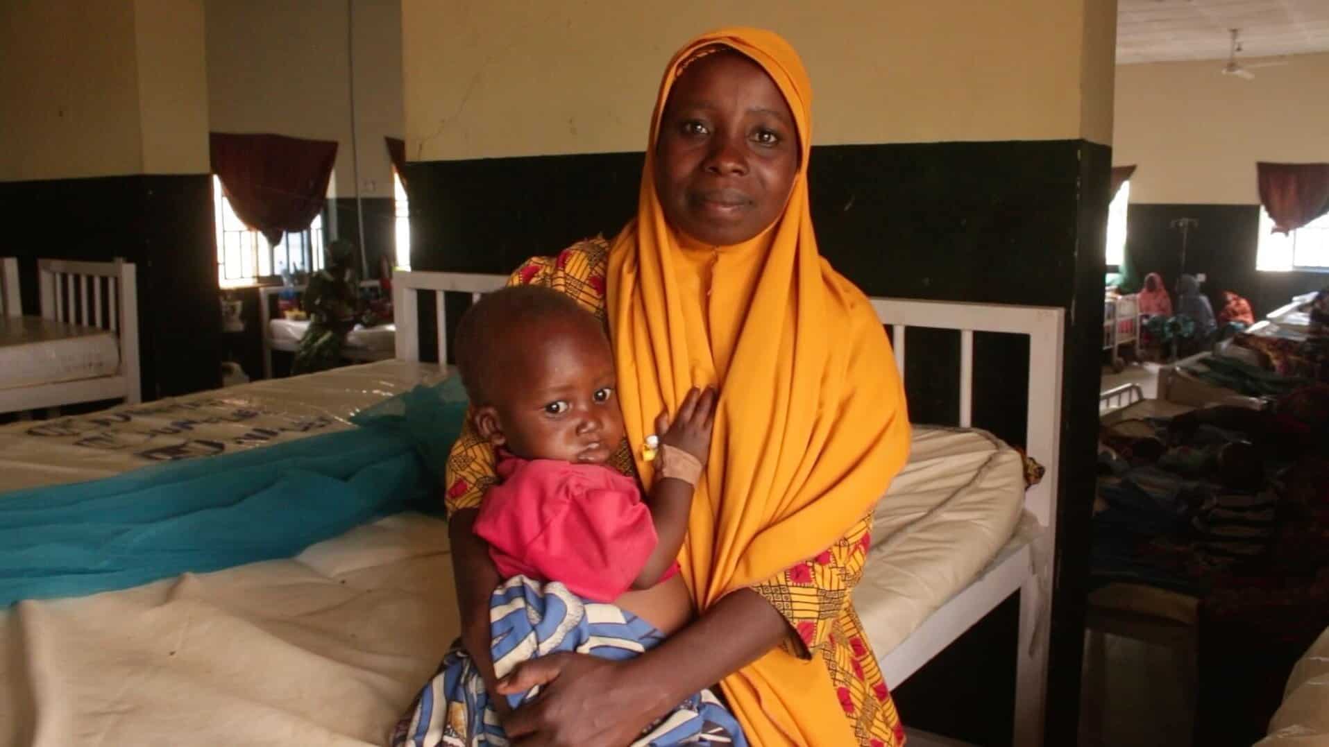 A woman with a headscarf smiles at the camera as she holds a young child in a
hospital room.