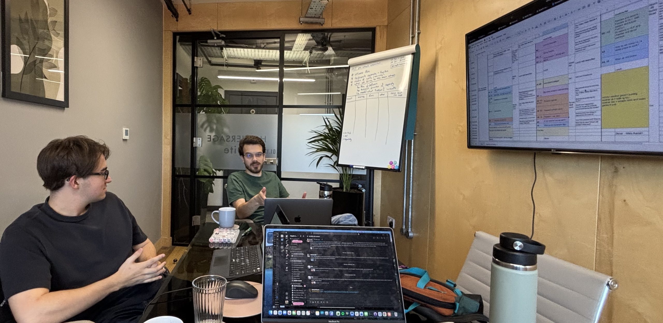 Two researchers talking across a meeting-room table, a handwritten research agenda on a flip chart behind them and a planning spreadsheet on the wall screen.
