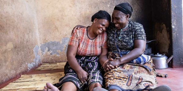 Two women sitting on a mat, smiling and laughing together as they hug.